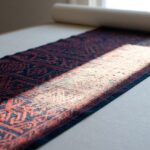 A patterned indigo and red textile laid flat on pale archival board, crossed by a hard-edged band of window daylight in which the dye has bleached to chalky pale, with a rolled blackout shade out of focus above.