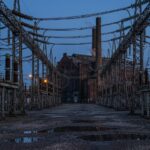 A high-voltage switchyard at dusk: rows of steel lattice towers and porcelain insulators, conductors sagging overhead, wet gravel and puddles in the foreground, and an old brick generating hall with a smoking stack behind.