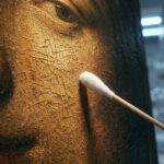 Extreme close-up of the cracked, golden varnished surface of an old oil painting, lit from the left, with the white cotton tip of a sampling swab resting against it; blurred laboratory glassware and cold lamps behind.