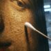 Extreme close-up of the cracked, golden varnished surface of an old oil painting, lit from the left, with the white cotton tip of a sampling swab resting against it; blurred laboratory glassware and cold lamps behind.