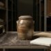 A single glazed stoneware storage jar and a small stack of blank paper on a museum handling cart in a quiet storage room.