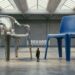 Two enormous stackable garden chairs, one polished steel and one matte blue, stand in an empty exhibition hall with a tiny figure looking up between them.