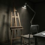 An empty wooden easel and an unoccupied metal chair under a single bright inspection lamp in a dark room, with white cotton gloves lying on a bare steel table.