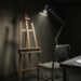 An empty wooden easel and an unoccupied metal chair under a single bright inspection lamp in a dark room, with white cotton gloves lying on a bare steel table.