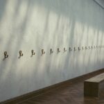 A long empty museum gallery wall in raking daylight, fitted with a single receding row of small bare brass picture hooks at even intervals; nothing hangs on them, there are no labels, and a plain wooden bench stands on the parquet floor.