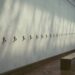 A long empty museum gallery wall in raking daylight, fitted with a single receding row of small bare brass picture hooks at even intervals; nothing hangs on them, there are no labels, and a plain wooden bench stands on the parquet floor.