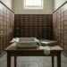 An empty archival storage box lined with acid-free tissue sits open on a plain table in a museum collections room, flanked by white cotton handling gloves, with rows of old wooden specimen drawers lining the walls under a high window.