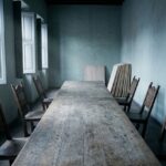 An empty guild meeting hall: a long bare oak table with carved wooden chairs pushed back at uneven angles, half-open shutters letting in cold daylight, and a stack of blank unfinished wooden painting panels leaning against the far wall.