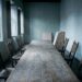 An empty guild meeting hall: a long bare oak table with carved wooden chairs pushed back at uneven angles, half-open shutters letting in cold daylight, and a stack of blank unfinished wooden painting panels leaning against the far wall.