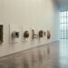 A long pale museum wall on which unlike objects — an old framed portrait, a white helmet, a ceramic jar, a wooden chair, a metal machine part, a folded textile — hang in one evenly spaced row at identical height, with no labels.