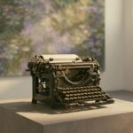 A worn antique typewriter alone on a plinth in a museum gallery, an out-of-focus impressionist garden painting in greens and violets on the wall behind it, warm window light, no people.