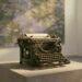 A worn antique typewriter alone on a plinth in a museum gallery, an out-of-focus impressionist garden painting in greens and violets on the wall behind it, warm window light, no people.