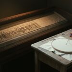 A museum display case holds the Bayeux Tapestry mid-scene, its embroidered figures stopping partway across the linen while the rest of the roll continues on, blank. Beside the case, a wooden table holds an empty embroidery hoop, a needle, and skeins of colored thread, as if someone has just stepped away to begin their own ending.