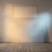 A large blank primed canvas leaning against a pale wall in an empty room, warm daylight from a window on the left and a cold spotlight from the right meeting across its surface, with a faint unfaded rectangle on the wall behind.