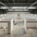 An empty convention-centre exhibition hall before an art fair opens: rows of identical bare white booth partitions on a wide concrete floor, one empty plinth, a rolled carpet, lighting trusses overhead.