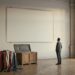 A tall empty official lobby wall with a large pale rectangle where a big painting has been removed, a foam-lined shipping crate and a flag-coloured cloth on a trolley below, one man looking up.