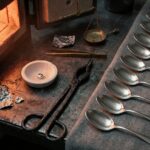 An assay bench beside an open furnace: a chalky porous cupel holding a single bright bead of metal, iron tongs, lead foil and a small brass balance pan, with a row of plain unmarked silver spoons laid out on grey cloth to the right.