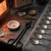 An assay bench beside an open furnace: a chalky porous cupel holding a single bright bead of metal, iron tongs, lead foil and a small brass balance pan, with a row of plain unmarked silver spoons laid out on grey cloth to the right.
