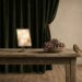 A dark green velvet curtain drawn back by a hand to show a blank wall, beside a wooden table with a bowl of grapes, a small tablet with a blurred glowing screen, and a sparrow perched at the table edge.