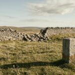An upland pasture in bright late-afternoon light: a long dry-stone wall running across the slope has collapsed into a wide breach, while in the foreground a short, plain, uncarved granite boundary post stands intact and upright, casting a long shadow across the grass.