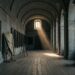 An empty vaulted granite gallery lit by one high window, large dark paintings on the wall, one on a trestle half uncovered from a dust sheet, a wooden stepladder beside it.
