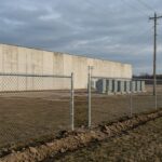 A new chain-link fence with freshly set posts in turned earth runs across the foreground of a cleared field; beyond it a long windowless pale concrete building and a row of grey transformer cabinets under an overcast sky.