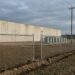 A new chain-link fence with freshly set posts in turned earth runs across the foreground of a cleared field; beyond it a long windowless pale concrete building and a row of grey transformer cabinets under an overcast sky.