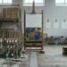A tall unfinished blue and gold panel on a wooden easel in an old workshop. The left side is fully stocked with pigment jars, brushes, stretcher frames and scaffolding; the right side is bare, with empty trestles, iron hooks and pale rectangles on the wall where things once hung.