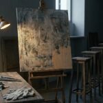 A dim studio at dusk: a large abstract painted panel on an easel, lit by a single lamp, its lower corner left as bare wood, with a row of empty wooden stands receding into shadow beside it.