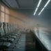 An empty corporate meeting room at dusk: a long bare table with a dense row of office chairs crowded along one side, a single chair alone opposite, and faint pale rings on the carpet where chairs have been removed.