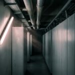 A dim machine hall of tall blank metal cabinets with overhead coolant pipes, lit by a single bare fluorescent tube mounted diagonally on the side wall like a light sculpture.