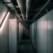 A dim machine hall of tall blank metal cabinets with overhead coolant pipes, lit by a single bare fluorescent tube mounted diagonally on the side wall like a light sculpture.
