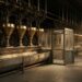 A long dim industrial sorting hall: a row of polished brass funnels feeding a continuous steel trough, with one small empty glass inspection booth standing open and unoccupied partway along the run.