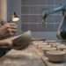 A potter's hands brushing the rim of a handmade stoneware bowl, with a row of slightly different bowls on the bench and an idle industrial robot arm beside a wall of identical grey tiles.
