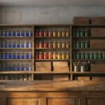 The interior of an old artists materials shop at closing time: a bare polished wooden counter, shelves of unlabelled glass jars of blue, red, yellow and green dry pigment, stacked plain wooden paint boxes, brushes in tin canisters, rolls of blank canvas, and bare plaster walls showing faint pale rectangles where pictures once hung.