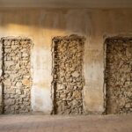 An empty Provencal salon with three tall rectangular voids cut out of the plaster wall, exposing bare stone and lath where painted panels were removed.